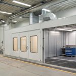 A large industrial paint booth with bright lighting, metal walls, and ventilation systems, showing safety equipment and workspace setup for automotive or machinery painting.