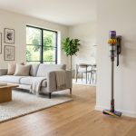 A Dyson cordless vacuum standing in a sunlit minimalist living room with hardwood floors.