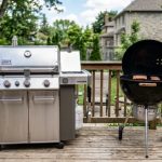 Stainless steel gas grill next to a black charcoal grill on a deck.